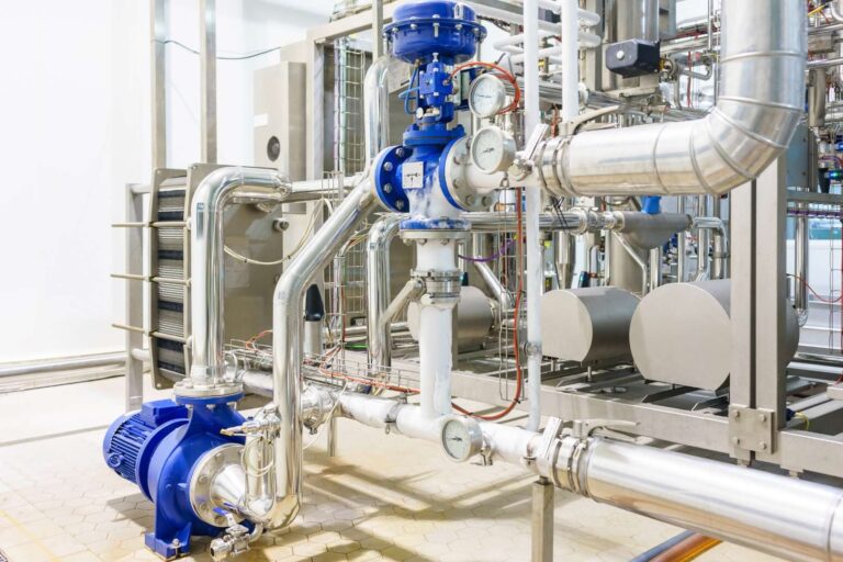 Sistema de tuberías industriales con una válvula azul y componentes de acero inoxidable, utilizado para el procesamiento de fluidos en una planta de fabricación. La instalación incluye manómetros y una bomba, lo que pone de relieve una ingeniería avanzada para operaciones eficientes.